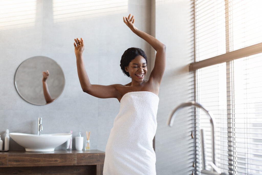 Cheerful young african american lady covered in white towel dancing and laughing in modern bathroom with window, enjoying morning sun light, taking bath, copy space. Skin and body care concept