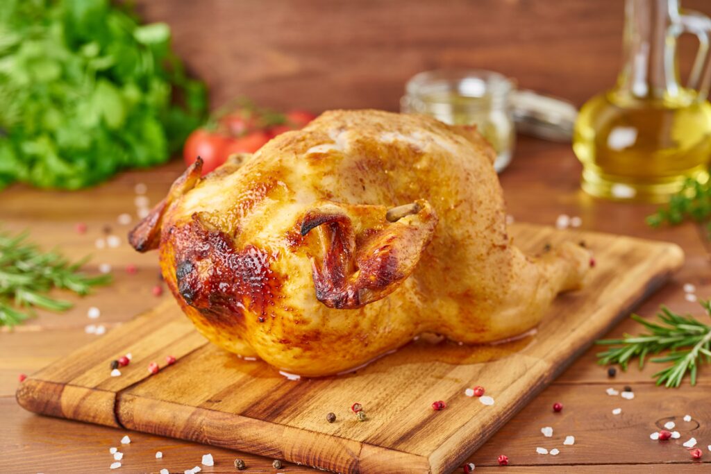 Whole roasted chicken on wooden cutting board on dark brown wooden table, side view